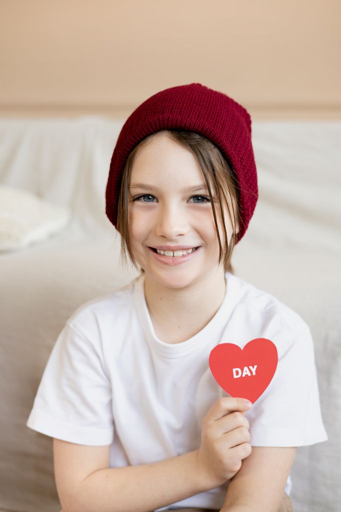 Child holding a red heart craft with 'DAY' sign indoors, smiling warmly.